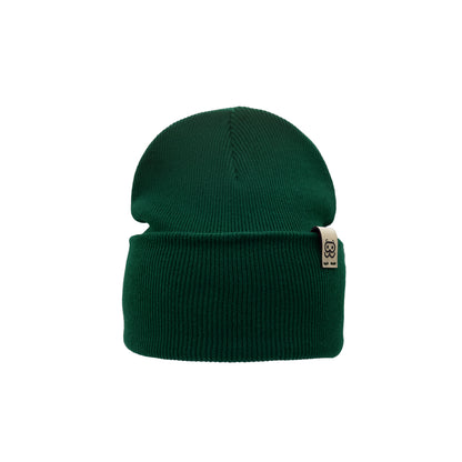 A green knit Organic Beanie from Goggleface with a folded cuff is shown. The beanie features a small rectangular fabric tag on the cuff with minimalistic branding. The texture of the knit is visible, giving it a cozy, warm appearance, making it a timeless accessory in sustainable fashion.
