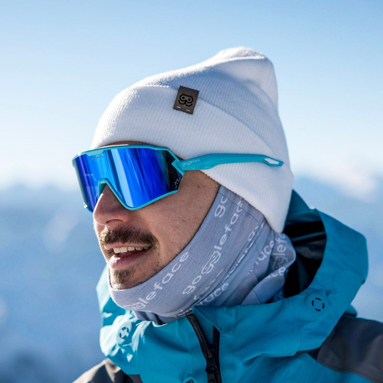 A person wearing a Goggleface Color Beanie made from recycled materials, reflective blue sunglasses, a grey face covering, and a blue winter jacket is smiling. The background is a snowy, mountainous landscape under a clear sky.