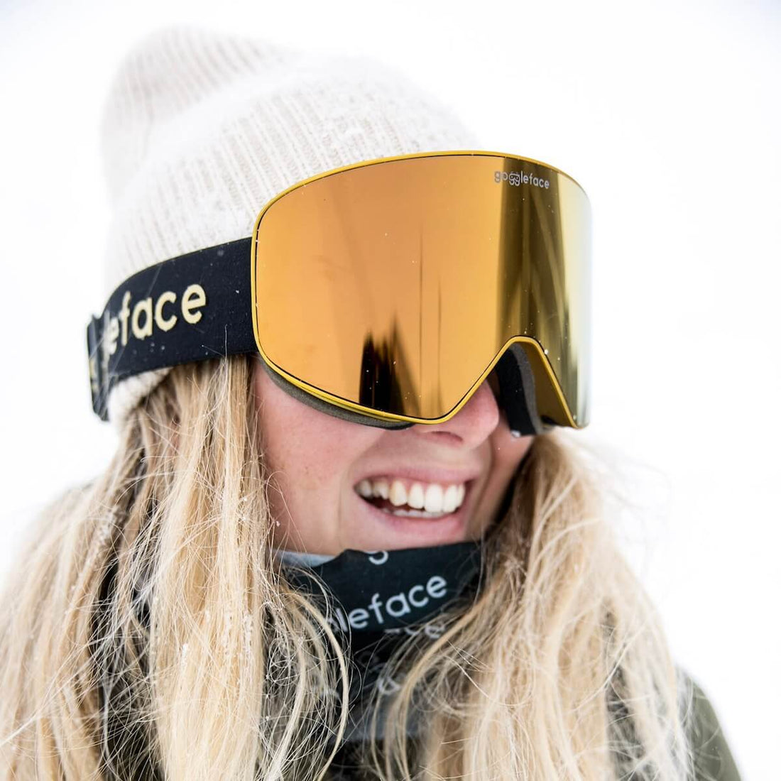 A person with long, blonde hair is smiling and wearing large, reflective orange ski goggles from the Goggleface Straight Line | Gold Rush collection and a knit beanie. Their neck is covered with a patterned neck gaiter. The background showcases a snowy, white landscape perfect for a winter adventure.
